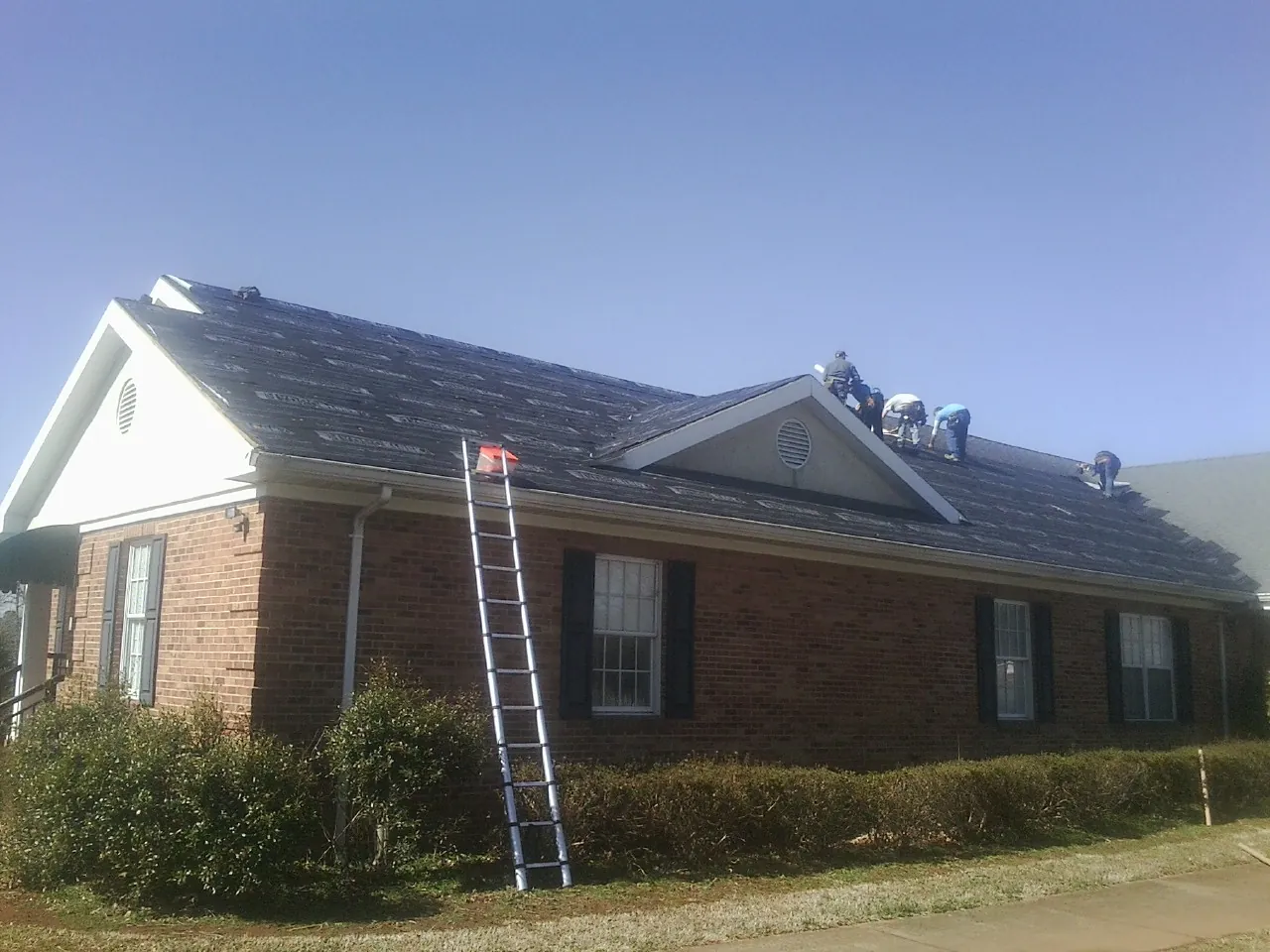 Crew tearing shingles off a brick ranch roof with a ladder set — Metal Roof Repair work by our crew in White River Junction