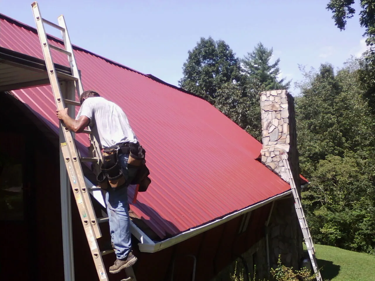 Roofer on a ladder beside a red metal roof and stone chimney — Soffit & Fascia Repair by Roofing Brothers in White River Junction